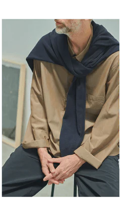 Lookbook de la collection SS25 de Still By Hand. Un homme porte une chemise beige et un pantalon foncé. Il a noué un pull autours de ses épaules.