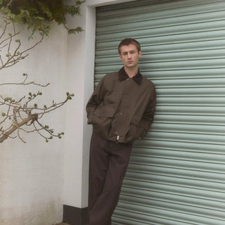 Un homme portant une veste Norse Project en suédine verte, un pull tricoté et un bonnet marron, se penchant légèrement dans un cadre minimaliste. Découvrez le style unique de la collection Norse Project, alliant élégance scandinave et confort.