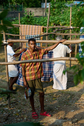 Un jeune homme posant fièrement près d'un étendage de tissu aux motifs à carreaux, incarnant le style décontracté de la collection de Original Madras Trading Company. L'image met en avant la qualité et l'authenticité des produits.