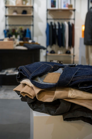 Un tas de vêtements présentés dans un magasin, avec en premier plan une veste en jean et plusieurs t-shirts et sweatshirts pliés au fond. Idéal pour compléter la collection Manteaux, Vestes & Gilets.