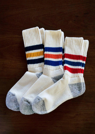 Chaussettes Rototo aux couleurs vives, avec des rayures bleues, rouges et jaunes, posées sur une surface en bois. Parfaites pour un look décontracté, ces chaussettes combinent style et confort typiques de la marque japonaise Rototo.