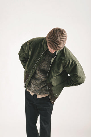 Un homme portant une veste Norse Project en suédine verte, un pull tricoté et un bonnet marron, se penchant légèrement dans un cadre minimaliste. Découvrez le style unique de la collection Norse Project, alliant élégance scandinave et confort.