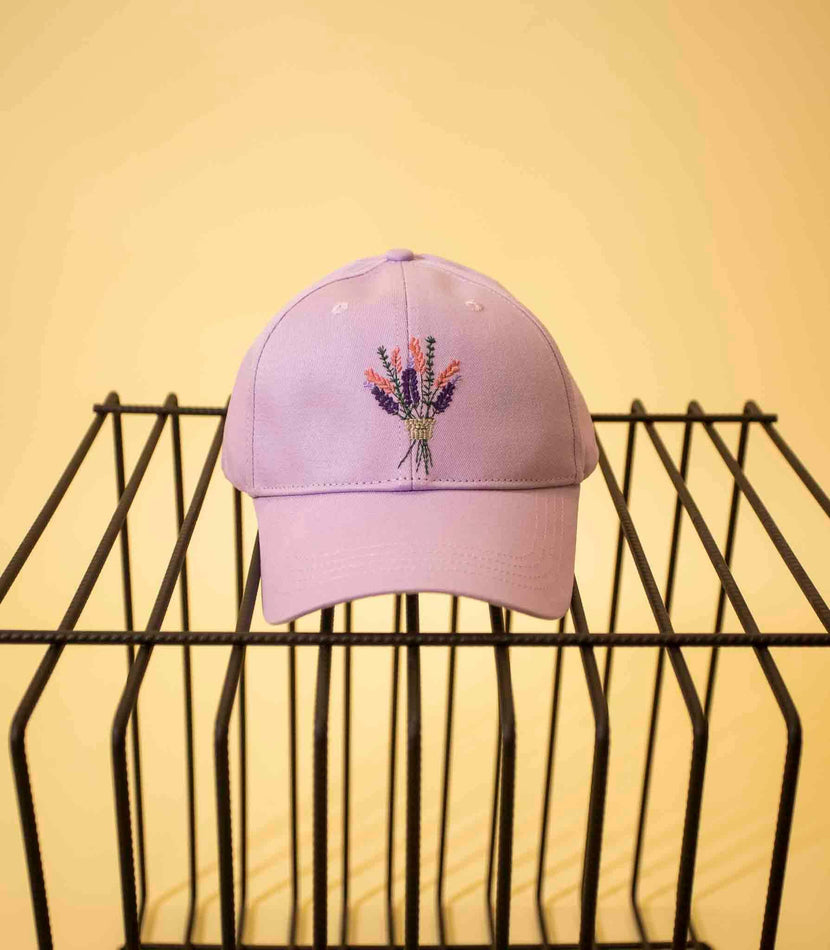 Casquette bouquet violet de Caleb photographiée sur un support noir avec un fond jaune. La casquette est lilas et présente une broderie colorée représentant un bouquet de fleurs.