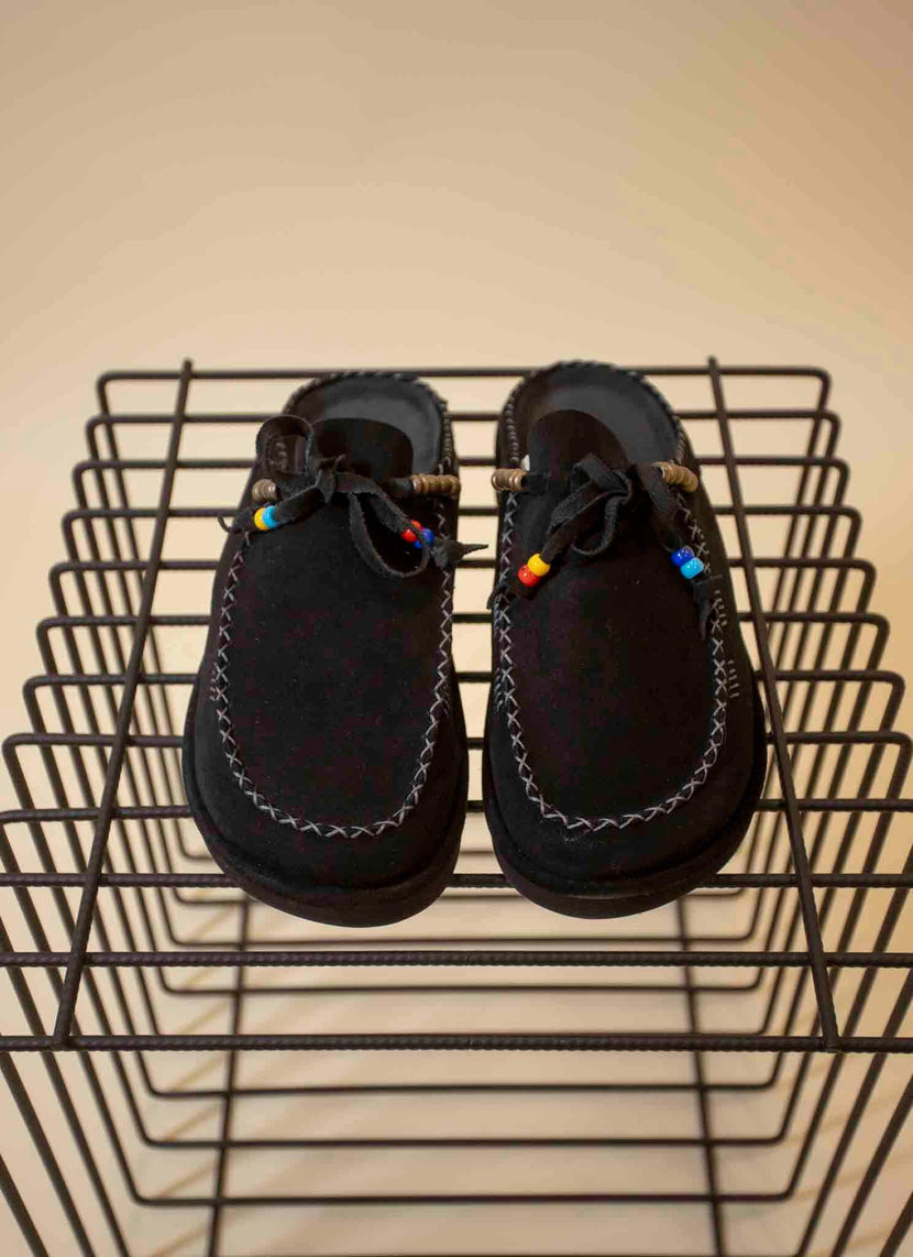 Chaussures noires KAIA de Caleb posées sur une étagère en métal, avec des détails colorés sur les lacets.